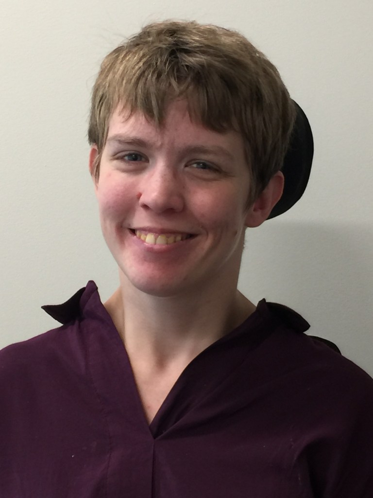 Headshot of Miriam Spies. White woman, smiling face, blue eyes, short blondish hair, against black wheelchair headrest.